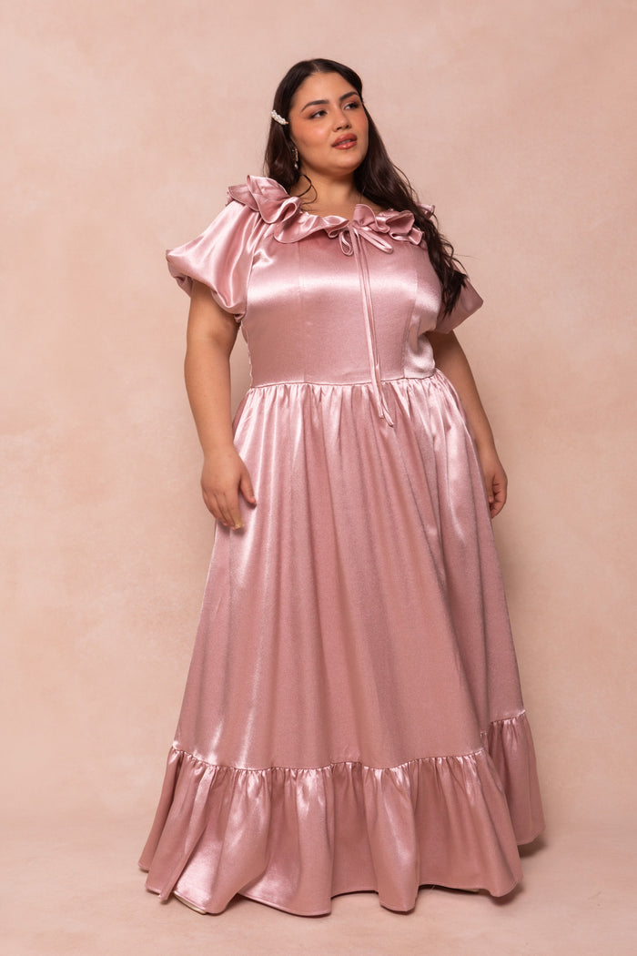 A woman stands against a beige background in the Billie Dress in Blush Satin by Ivy City Co, featuring short puff sleeves, a ruffled neckline with bow, and a flowing ruffled hem—perfect as a bridesmaid dress.