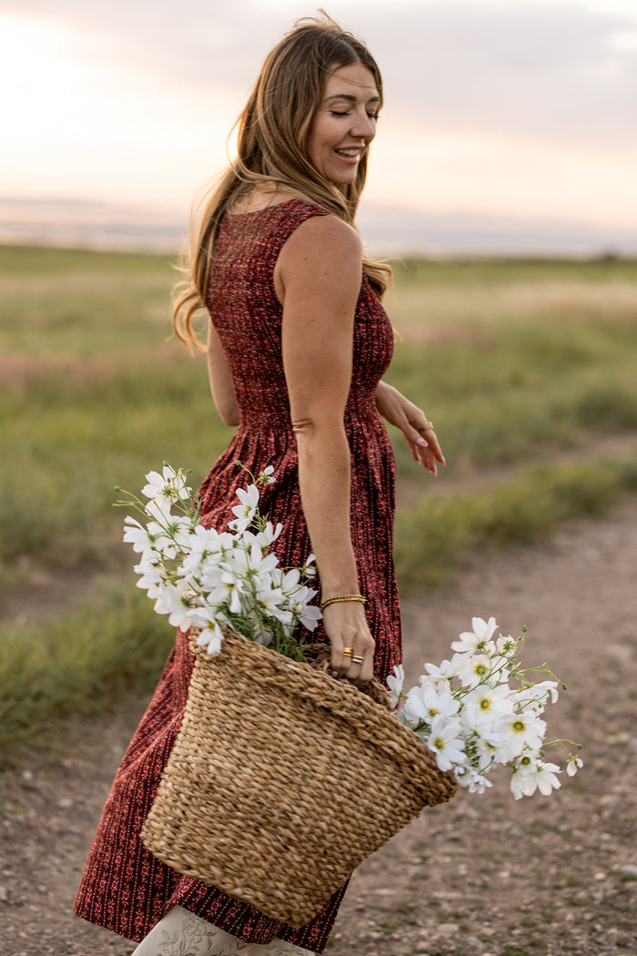 A woman in the Ana Dress in Sequoia Felicity Floral by Ivy City Co strolls outdoors on a dirt path, smiling and carrying a woven basket of white flowers. Green fields and a cloudy sunset sky complete the peaceful scene.