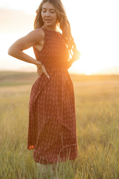 A woman wears the Ana Dress in Sequoia Felicity Floral by Ivy City Co, standing with a smile and eyes closed in a sunlit grassy field at sunset, one hand on her hip.