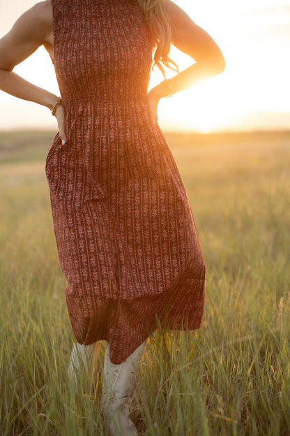 A woman wearing the Ivy City Co Ana Dress in Sequoia Felicity Floral stands hands-on-hips in a sunlit grassy field at sunset; her head is out of frame and golden light bathes the scene.