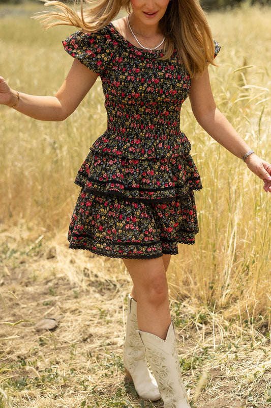 A woman in the Annalise Top in Obsidian Vintage Poppies by Ivy City Co walks through a sunlit field of tall, dry grass. Her face is partially out of frame, and her loose hair flows as she moves.