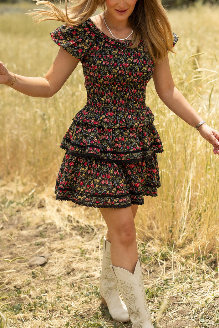 A woman wearing the Annalise Top in Obsidian Vintage Poppies by Ivy City Co with white cowboy boots walks through a grassy field, her arms slightly outstretched and her face partially out of frame.