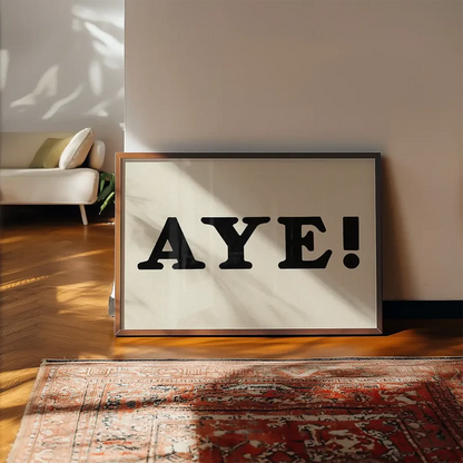 Aye Typography Poster Print framed, leaning on a wooden floor beside a rug