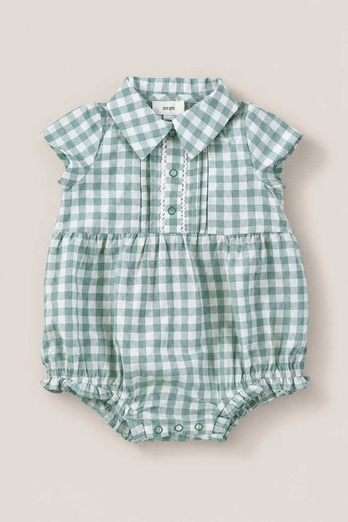 The Ivy City Co Baby Amber Romper in Sage Gingham features green and white cotton, short sleeves, a collar, button placket, and gathered leg openings. The vintage-inspired romper is shown laid flat on a neutral background.