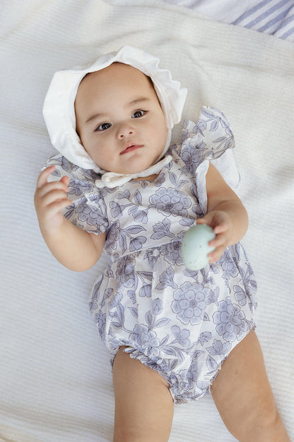 An infant dressed in the Ivy City Co Baby Hattie Romper in Blue Floral and a white bonnet lies on a soft white blanket, holding a small blue egg-shaped object in one hand and gazing upward with a calm expression.