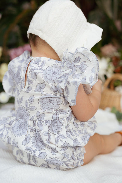 A baby dressed in the Ivy City Co Baby Hattie Romper in Blue Floral and a white bonnet sits on a white blanket, facing away, with a picnic basket and greenery softly blurred in the background.