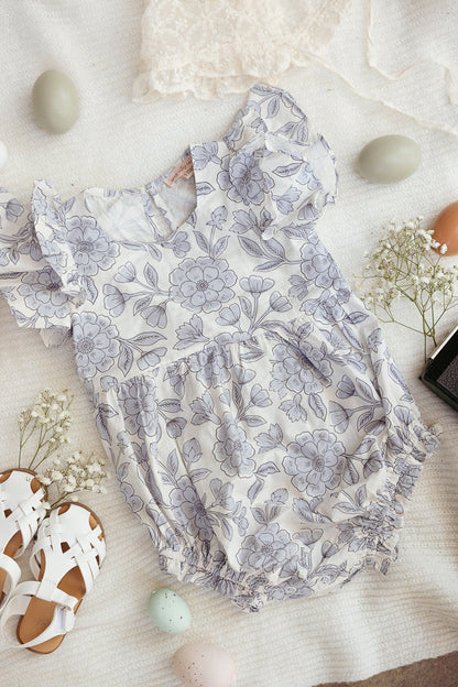 The Baby Hattie Romper in Blue Floral by Ivy City Co is displayed on a white blanket, accented with ruffled sleeves and surrounded by pastel Easter eggs, white sandals, delicate white flowers, and a lace cloth.