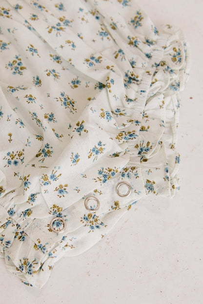 Close-up of Ivy City Cos Baby Madeline Romper in Blue, a white floral romper with blue and yellow flowers, ruffles, puff sleeves, and silver snap buttons—ideal for babies as a cute outfit or for special occasions.