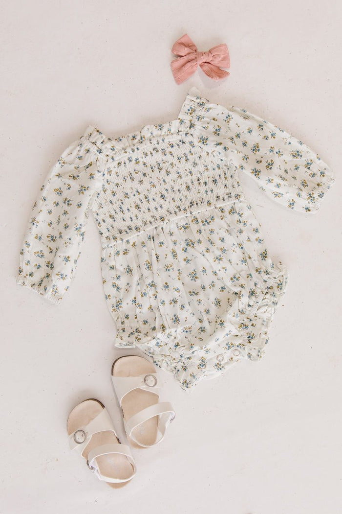 Flat lay of a sweet baby Easter outfit featuring the Ivy City Co Baby Madeline Romper in Blue, matching sandals with button details, and a pink fabric bow, all arranged on a light background.