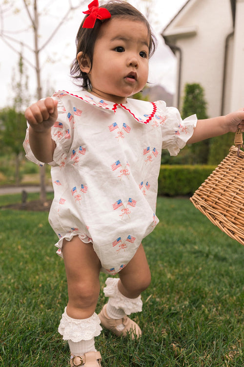 A toddler wears the Ivy City Co Baby Maisy Romper in Star Spangled, paired with white lace socks, beige shoes, a red bow, and holds a wicker basket on grass. Small American flags are featured; a house and trees appear in the background.