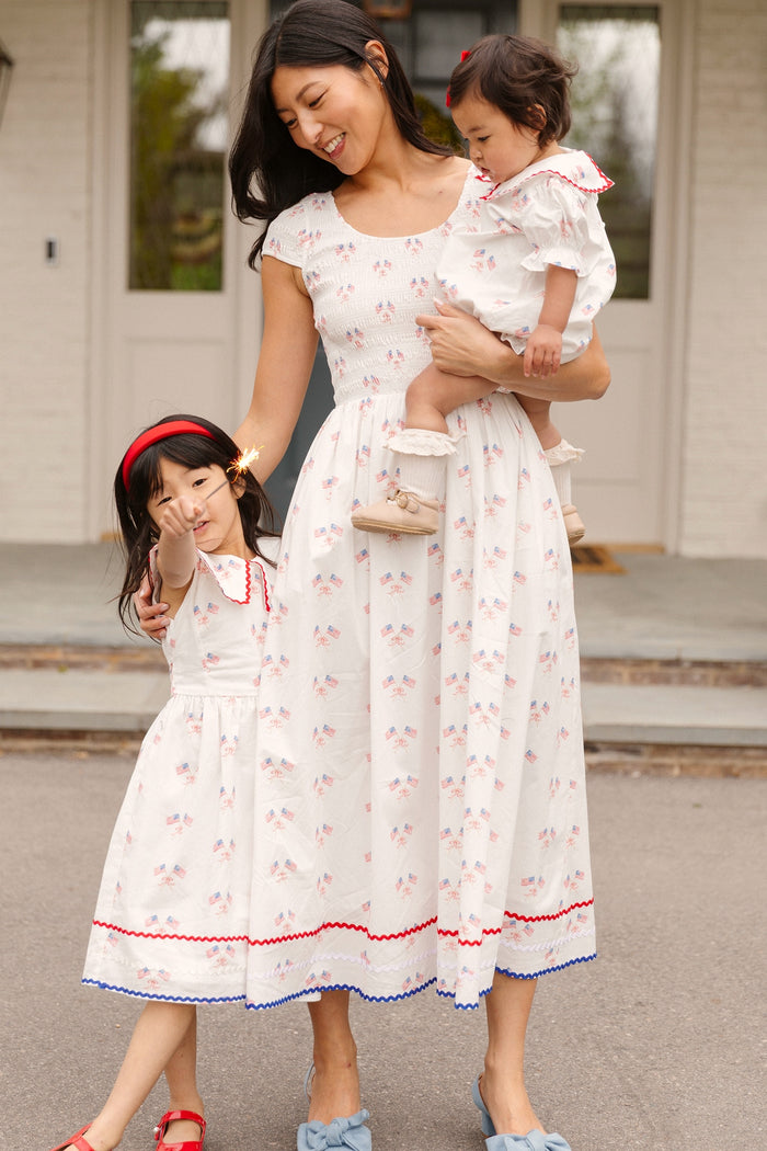 A woman and two girls stand outside in matching white dresses with red and blue details. The baby looks adorable in her Ivy City Co Baby Maisy Romper in Star Spangled as the woman smiles and holds her. One girl holds a sparkler.