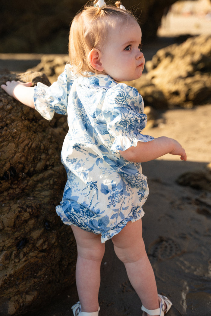 A toddler in Ivy City Co’s Baby Sophia Romper in Blue Floral and white sandals stands on a sunny beach near a large rock, sunlight highlighting the outfit and rocky background as the child looks over their shoulder.