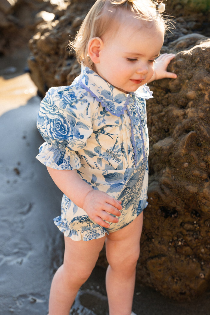 A toddler in the Ivy City Co Baby Sophia Romper in Blue Floral stands by a large rock on a sandy beach, gently touching it as sunlight highlights the romper’s delicate floral pattern.