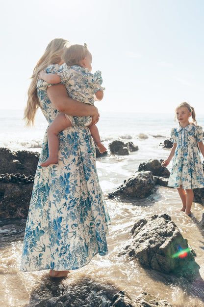 A woman stands barefoot on rocky sand by the ocean, holding a baby in the Ivy City Co Baby Sophia Romper in Blue Floral. Both wear matching blue floral dresses as a young girl in a similar dress looks on, sunlight highlighting the scene.