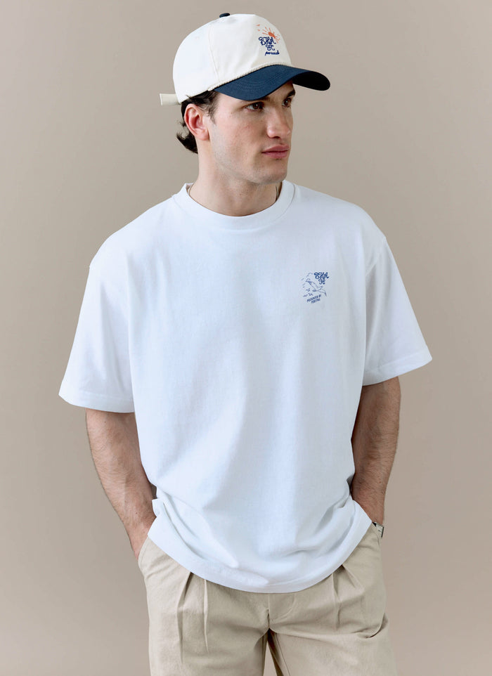 A model wears the white Beach Front T Shirt from the Percival x Brighton & Hove Albion F.C. collaboration, styled with a cap.