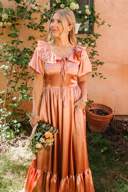 A woman wears the Ivy City Co Billie Dress in Apricot Crush Satin, featuring ruffled sleeves, as she stands outdoors with a bouquet of yellow-orange roses, surrounded by greenery and flowers against a peach wall.