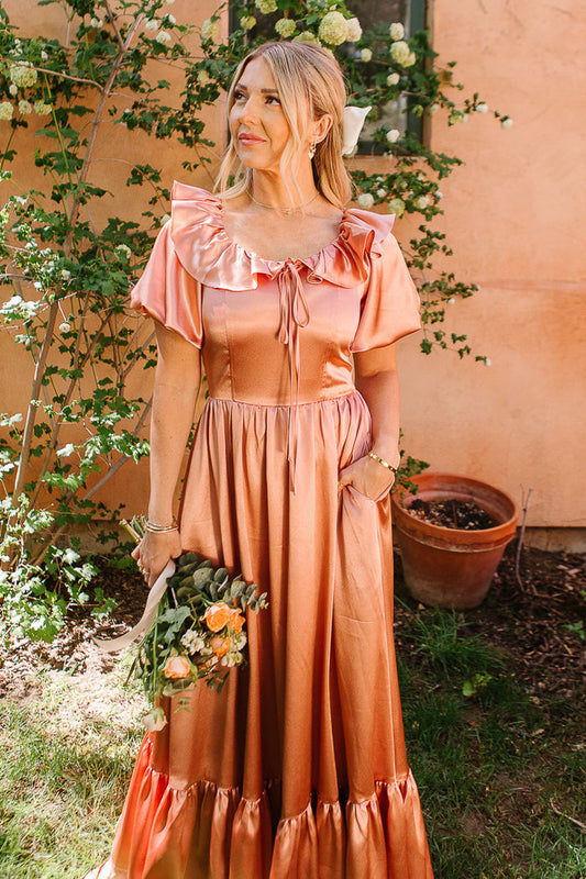 A woman wears the Ivy City Co Billie Dress in Apricot Crush Satin, featuring ruffled sleeves, as she stands outdoors with a bouquet of yellow-orange roses, surrounded by greenery and flowers against a peach wall.