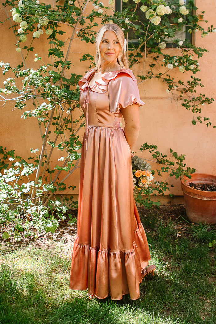 A woman in the Ivy City Co Billie Dress in Apricot Crush Satin stands on grass by green plants and a peach wall, holding flowers and smiling—an ideal satin bridesmaid dress with a shiny peach-pink glow.
