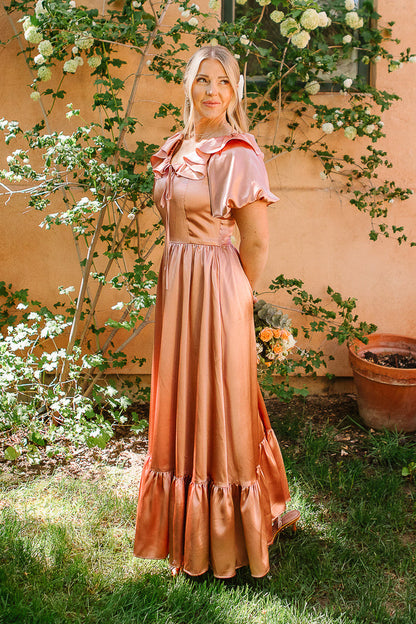 A woman in the Ivy City Co Billie Dress in Apricot Crush Satin stands on grass by green plants and a peach wall, holding flowers and smiling—an ideal satin bridesmaid dress with a shiny peach-pink glow.