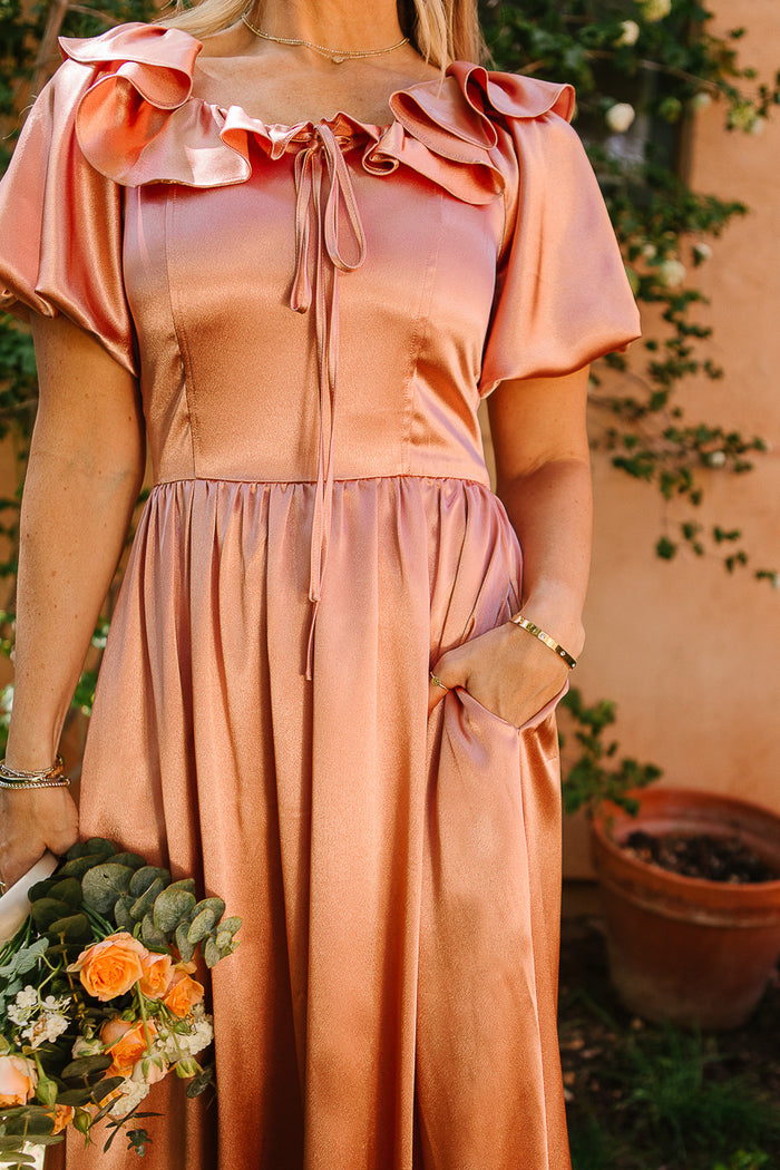 A woman in the Ivy City Co Billie Dress in Apricot Crush Satin stands outdoors holding orange and yellow flowers. The dress features ruffled sleeves, a gathered waist, and pockets. Her face is out of view.