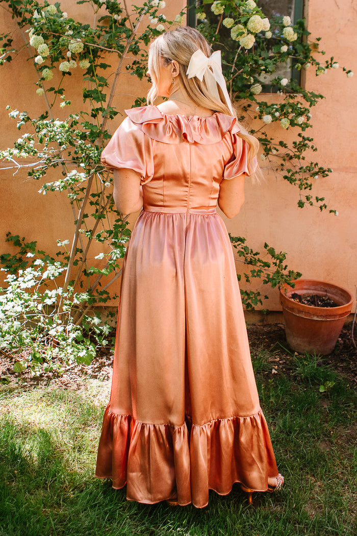 A woman with blonde hair tied in a white ribbon stands outdoors with her back to the camera, wearing the Ivy City Co Billie Dress in Apricot Crush Satin near a peach wall and green leafy plants.
