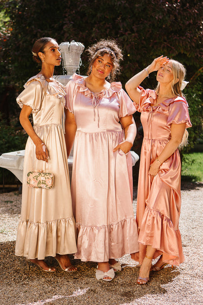 Three women pose outdoors in front of a fountain, smiling in flowy satin bridal party dresses from Ivy City Co—including the Billie Dress in Apricot Crush Satin—with greenery visible in the background.