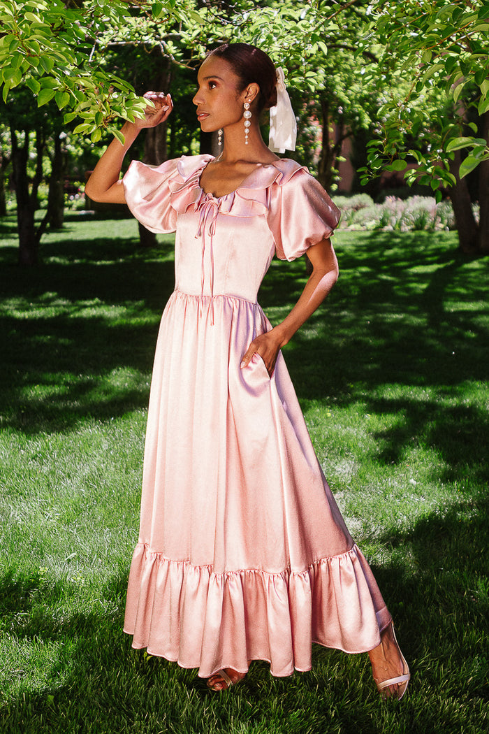 A woman stands on green grass, wearing the Ivy City Co Billie Dress in Blush Satin with ruffled short sleeves. She has a white ribbon in her hair and poses gracefully under leafy trees in bright sunlight.