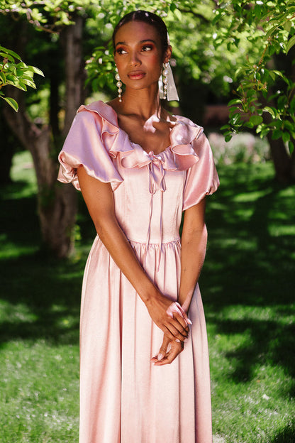 A woman stands outdoors in sunlight wearing the Billie Dress in Blush Satin by Ivy City Co, featuring ruffled sleeves and neckline. She has braided hair, long earrings, and is surrounded by green foliage.