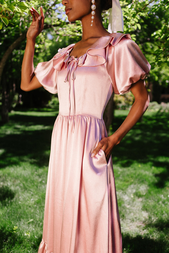 A woman stands outdoors in sunlight wearing the Ivy City Co Billie Dress in Blush Satin, featuring puff sleeves, a ruffled neckline, and pockets. She accessorizes with large pearl earrings and a hair ribbon; green trees and grass are behind her.