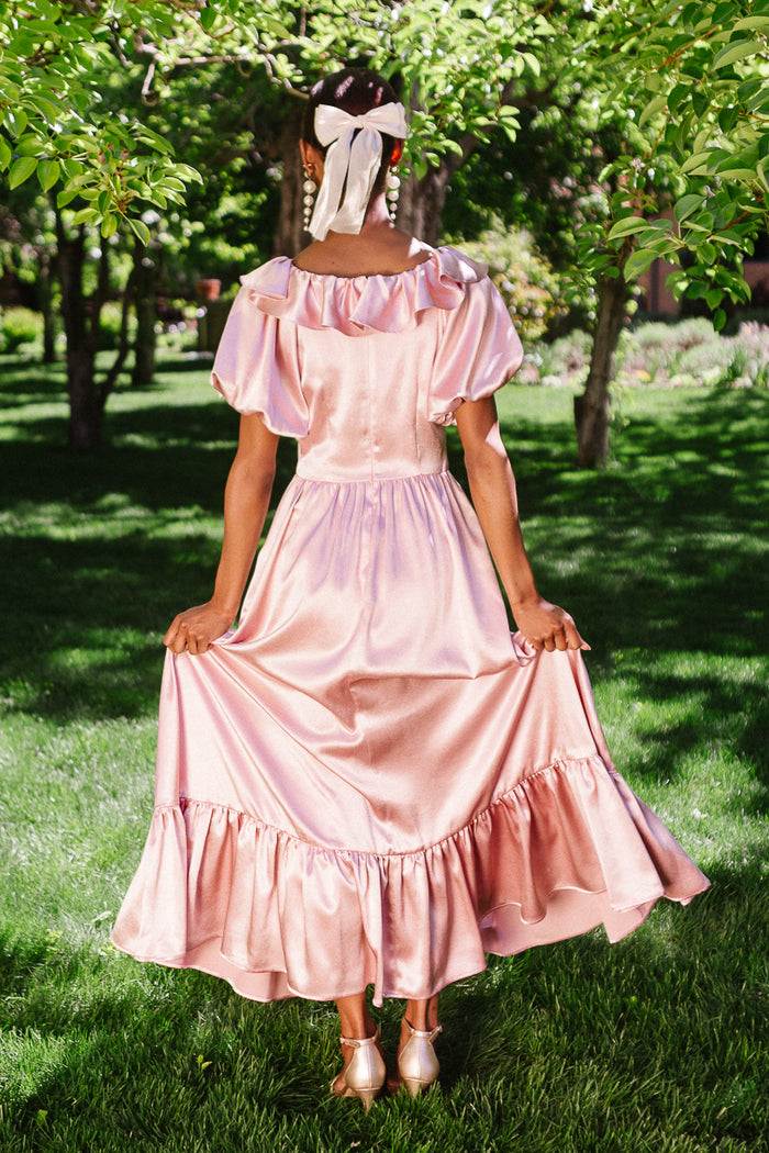 A person stands on grass, facing away, holding the sides of their Ivy City Co Billie Dress in Blush Satin. They wear shiny shoes and a white ribbon in their hair, surrounded by green trees with sunlight filtering through the leaves.