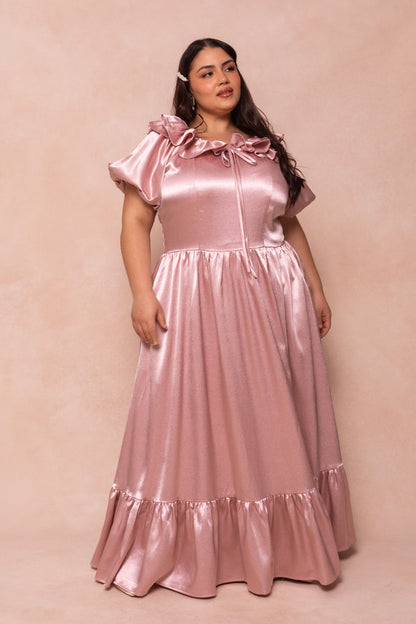 A woman stands against a beige background in the Billie Dress in Blush Satin by Ivy City Co, featuring short puff sleeves, a ruffled neckline with bow, and a flowing ruffled hem—perfect as a bridesmaid dress.