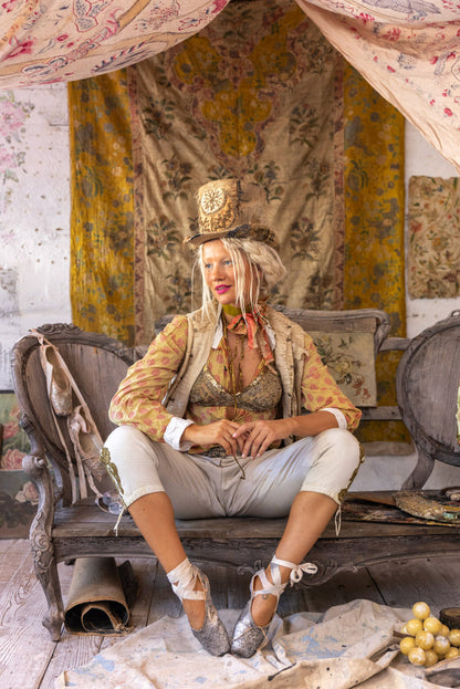 Woman in whimsical Magnolia Pearl artisan clothing, vintage top hat, and ballet shoes, seated on antique bench amid eclectic boho decor