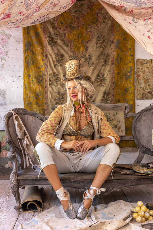 Woman in whimsical Magnolia Pearl artisan clothing, vintage top hat, and ballet shoes, seated on antique bench amid eclectic boho decor