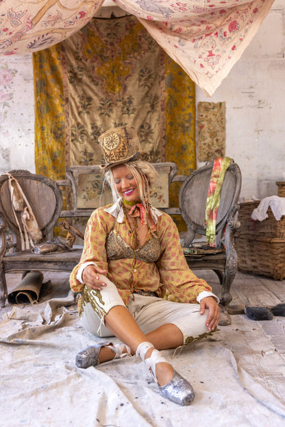 Woman in Magnolia Pearl artisan vintage clothing, whimsical top hat, and sequin shoes, seated in rustic boho room