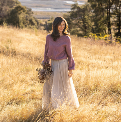 A woman stands in a sunlit field of dry golden grass, wearing a dusty mauve knit sweater and a long cream tulle skirt, holding a small bouquet of dried flowers.