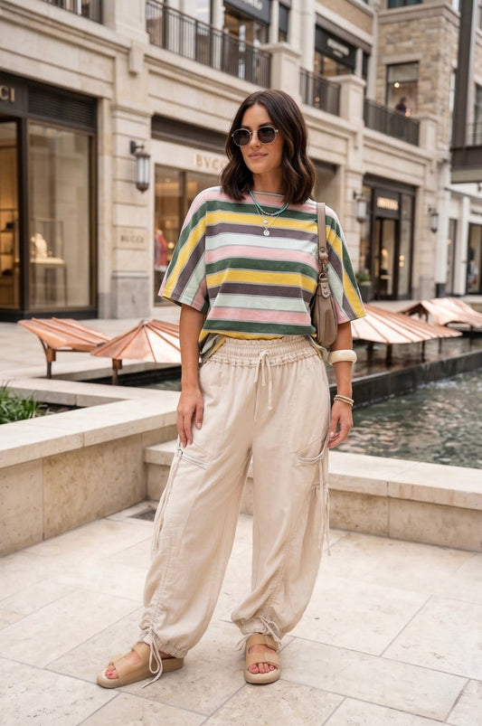 Soft striped oversized boutique tee in muted pastel tones featuring olive green, blush pink, mustard yellow, taupe, and cream stripes styled with neutral drawstring cargo pants for an effortless summer outfit aesthetic.