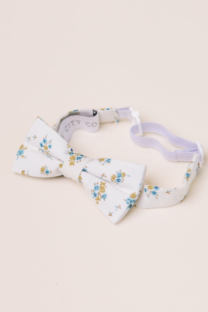 The Ivy City Co Ties in Madeline Blue Floral have a white base with blue and yellow blooms, an adjustable strap, and are great for family matching. Shown on a light beige background.