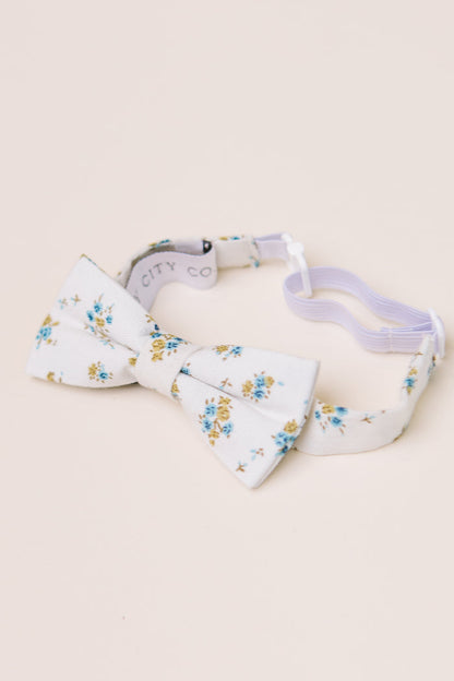 The Ivy City Co Ties in Madeline Blue Floral have a white base with blue and yellow blooms, an adjustable strap, and are great for family matching. Shown on a light beige background.