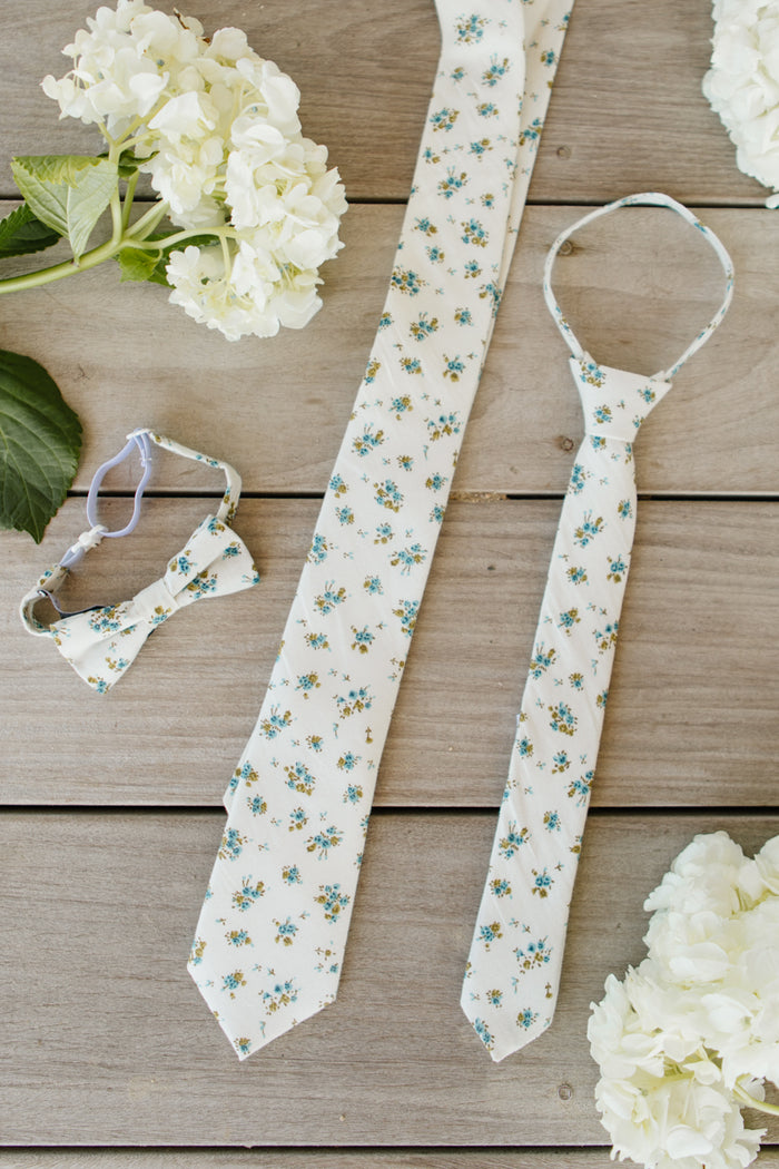 Ivy City Co’s Ties in Madeline Blue Floral—a necktie, child’s tie, and boys bow tie—are arranged on wood with white hydrangeas and green leaves for an adorable family matching look.