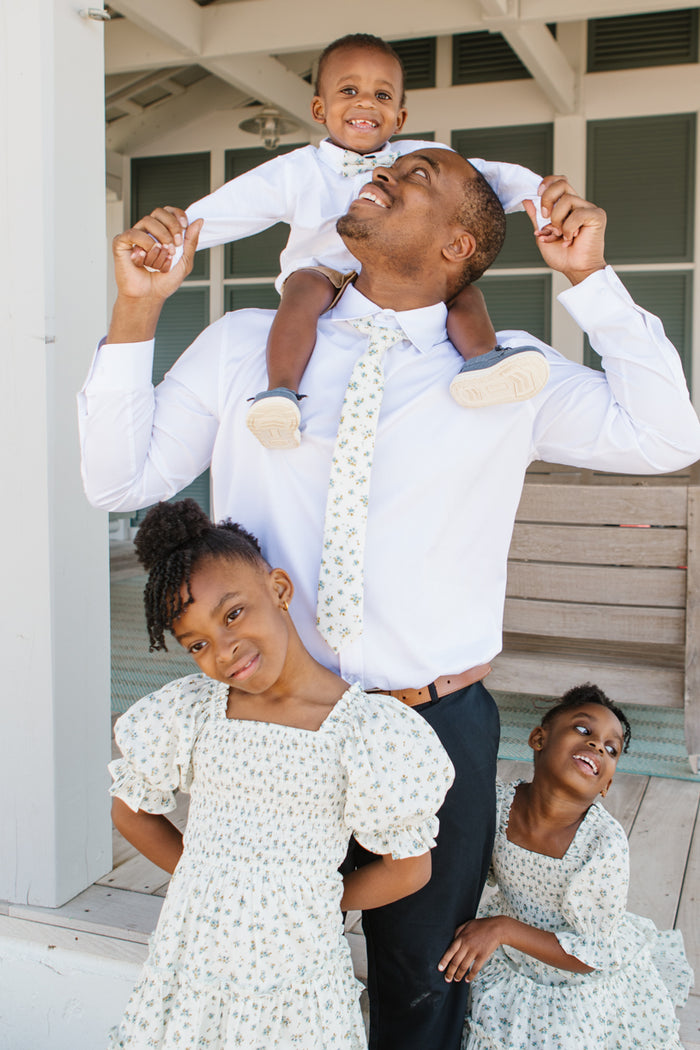 A man in a white shirt and Ivy City Co Ties in Madeline Blue Floral smiles while carrying a boy with a bow tie on his shoulders. In front, two girls in matching floral dresses pose—they all beam happily outside a building.