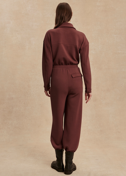 Woman turned around wearing a brown jumpsuit with black boots in front of a tan background