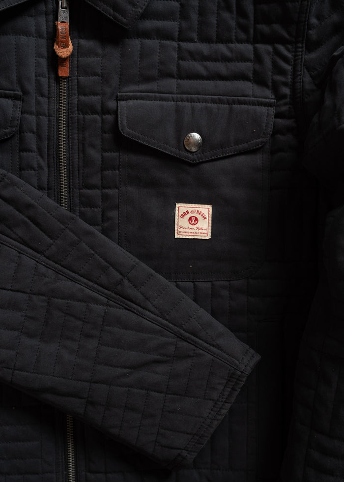 Close-up of a black quilted jacket with a front pocket, metal snap, zipper, and a small red and white "Iron Heart" patch.