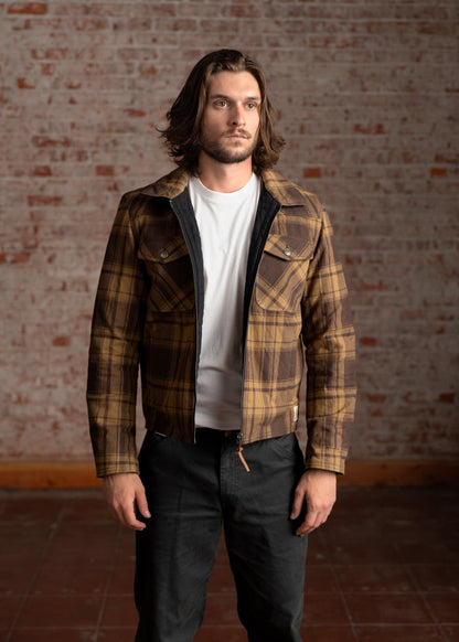 Man in a plaid jacket and white shirt stands against a brick wall, looking forward. Casual, stylish, and confident pose.