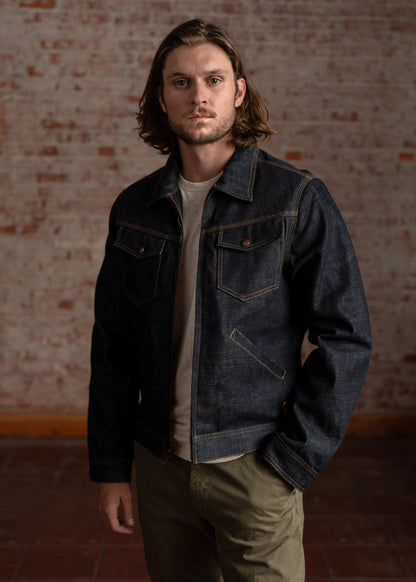 Man with long hair in denim jacket and khaki pants stands against a brick wall, looking directly at the camera.