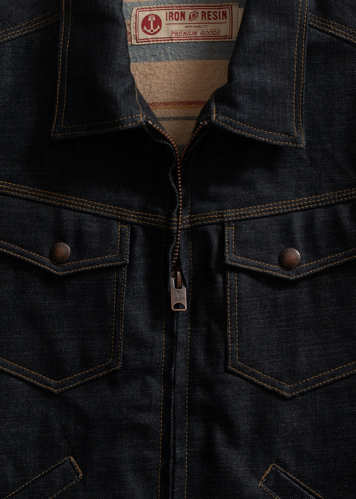 Close-up of a dark denim jacket with a zipper and two chest pockets. Label reads "Iron and Resin Premium Goods."