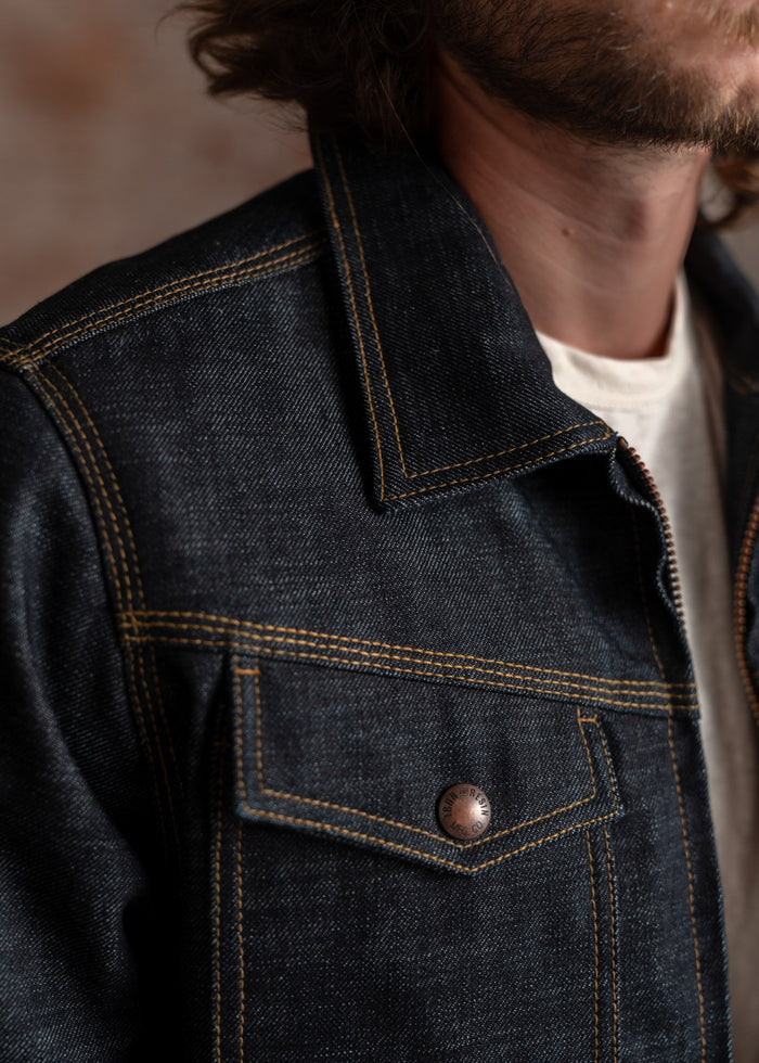 Close-up of a denim jacket with contrast stitching, highlighting a buttoned pocket and collar, worn over a white shirt.