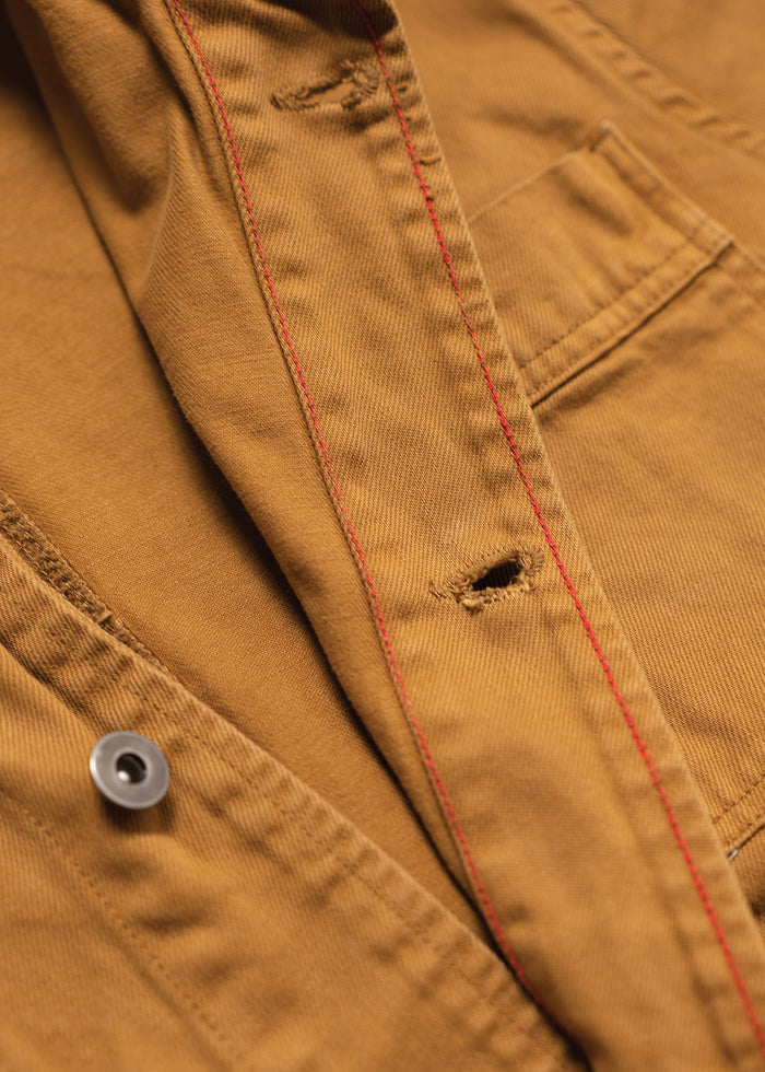 Close-up of a brown jacket with red stitching, showing a button and pocket detail. The fabric texture is clearly visible.