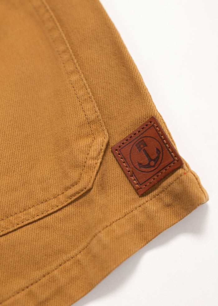 Close-up of a brown fabric corner with a stitched seam and a leather patch featuring an anchor logo, set against a white background.