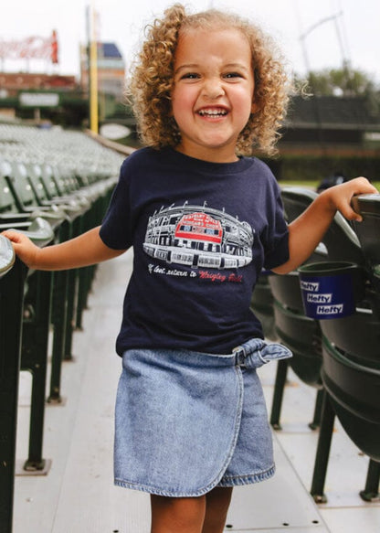 Child wearing a navy t-shirt with a graphic design and denim skirt standing in stadium seats.